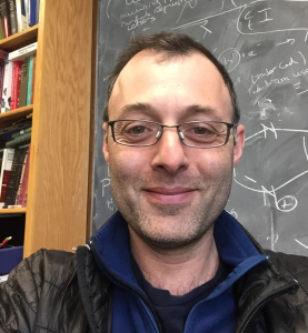 Headshot of a person wearing glasses and a dark jacket, photographed in front of a chalkboard covered with scientific equations and diagrams.