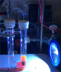 Two glass reaction vessels connected by a small horizontal tube are mounted in a laboratory apparatus. Wires and metal clips extend into the vessels through white stoppers. A bright blue lamp shines on the setup, illuminating colored solutions inside the tubes.