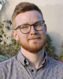Portrait of a person with short hair, glasses, and a beard, wearing a patterned button-down shirt and standing outdoors in front of a softly blurred garden and stone wall.
