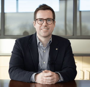 Portrait of a person wearing glasses, a checkered shirt, and a dark blazer, seated at a table in an office setting.